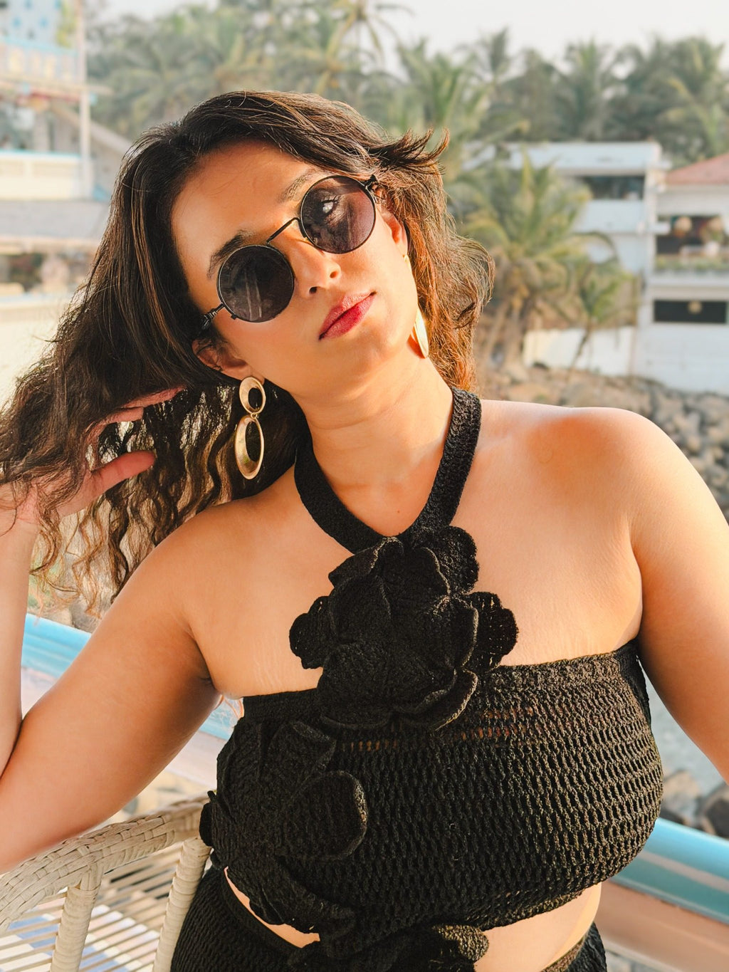 Close-up portrait of model in black crochet halter crop top with 3D floral appliqué, styled with round sunglasses and gold hoop earrings.