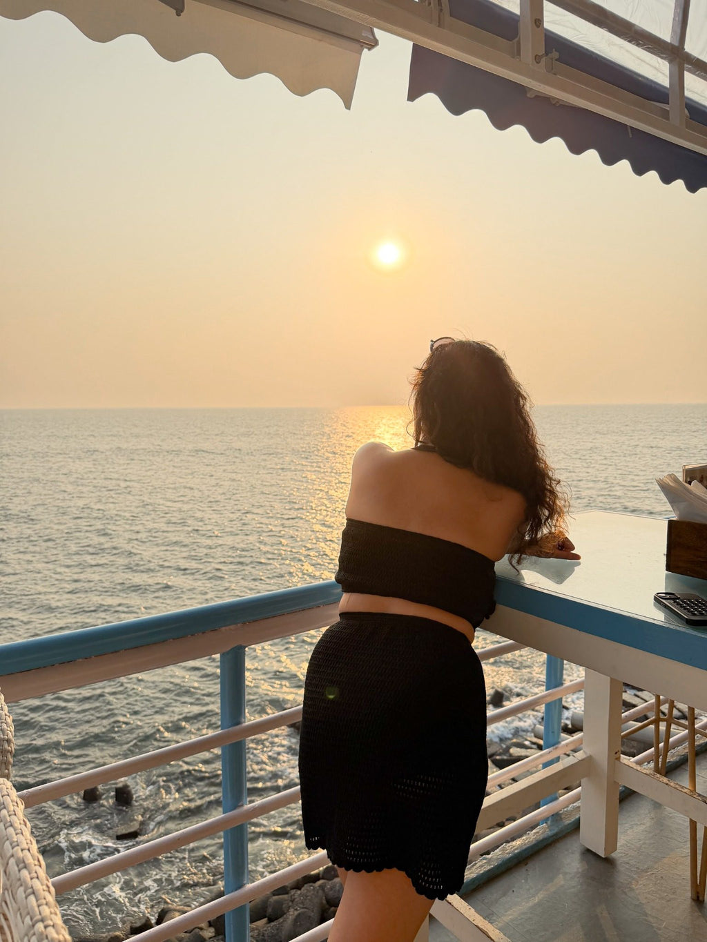 Back view of black crochet co-ord set with strapless back and crochet mini skirt, model looking at ocean sunset from balcony.